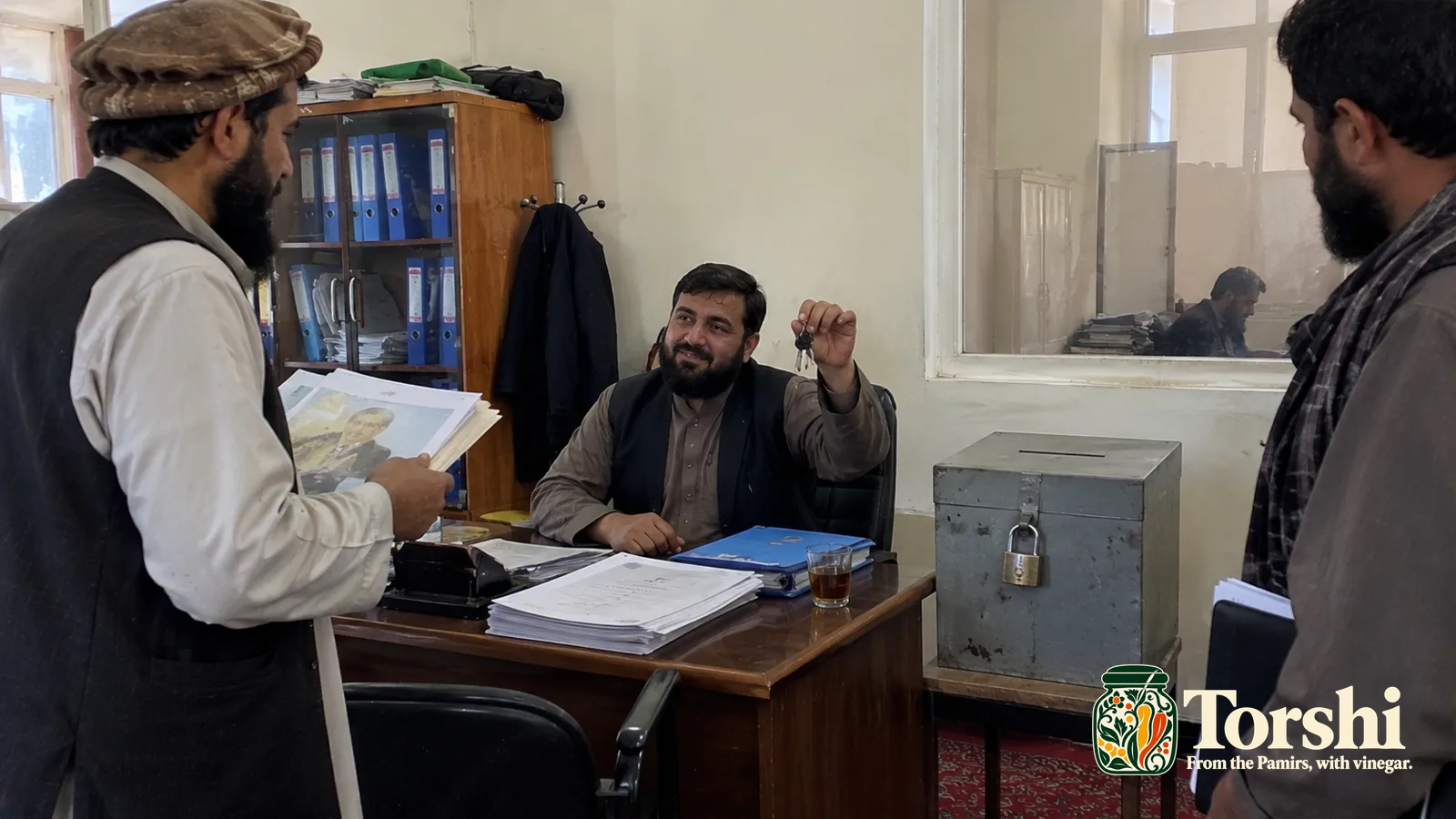 A seated man holds a key beside a locked complaints box on an office desk.