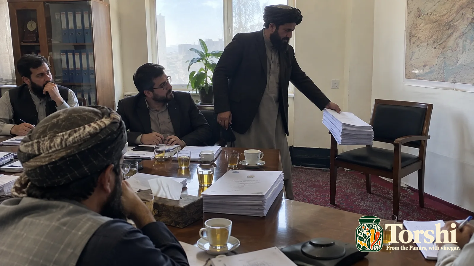 Men sit around a meeting table covered with papers and tea glasses.